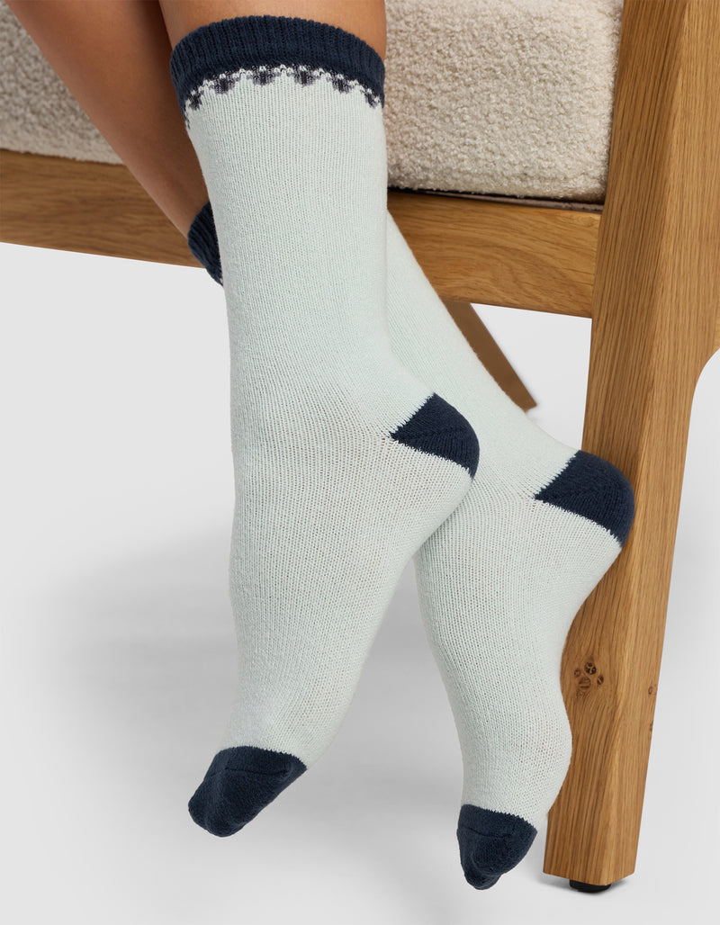 Close-up of a person wearing Cozy Earth's The Mistletoe Plush Lounge Sock, featuring light fabric with dark heels, toes, and cuffs, while sitting on a wooden chair with crossed feet on a neutral background.