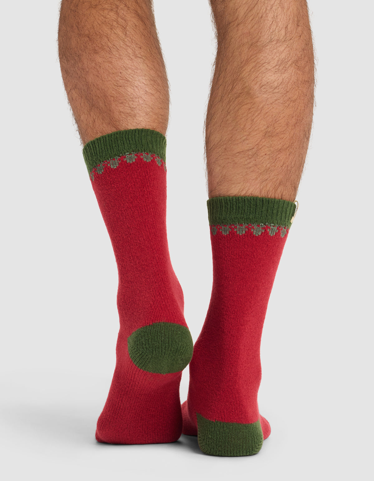 Close-up of a person's legs wearing The Mistletoe Plush Lounge Sock by Cozy Earth—red socks with green heels, toes, and cuffs—standing on a plain light background.