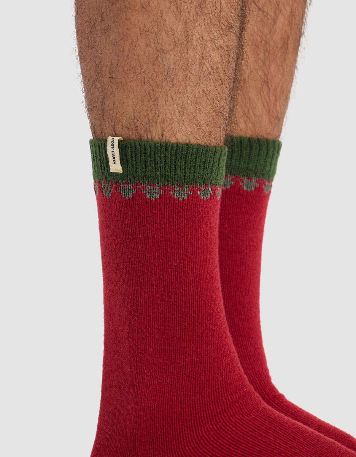 Close-up of a person's lower legs wearing The Mistletoe Plush Lounge Sock by Cozy Earth—red socks with green ribbed cuffs and a small white fabric tag—against a plain light background.