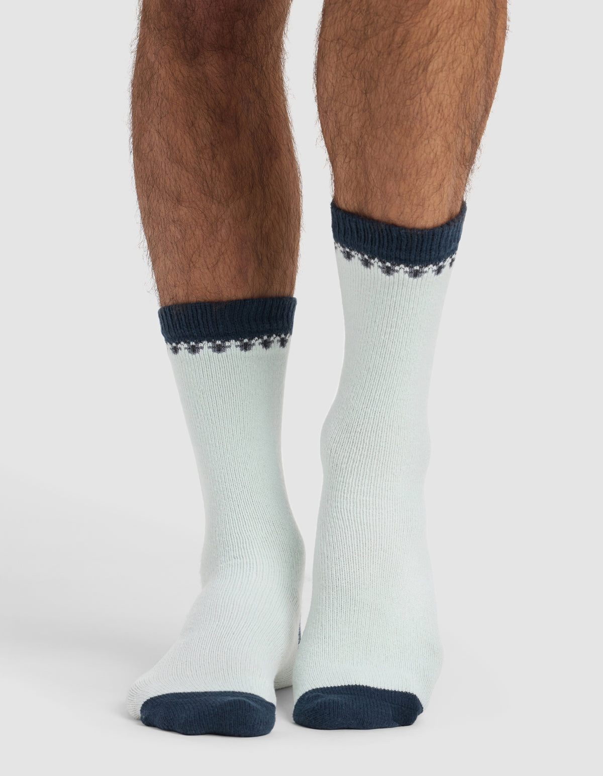 A person wears The Mistletoe Plush Lounge Sock by Cozy Earth: light-colored with dark blue toes, heels, cuffs, and a patterned band near the top. The background is plain and light.