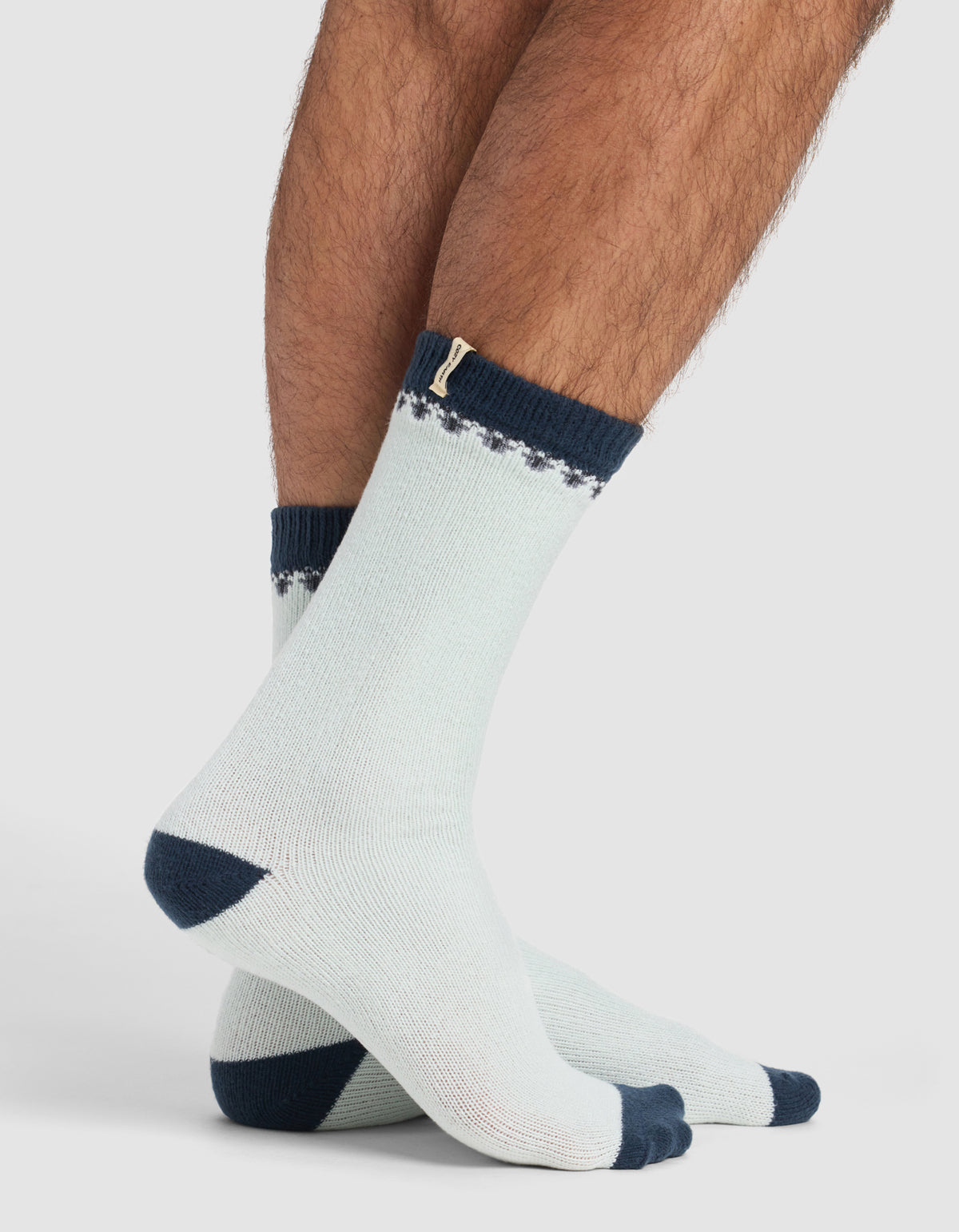 Someone stands on a white background wearing The Mistletoe Plush Lounge Sock by Cozy Earth, featuring light colors with dark blue toes, heels, and cuffs, plus a subtle decorative pattern near the top.