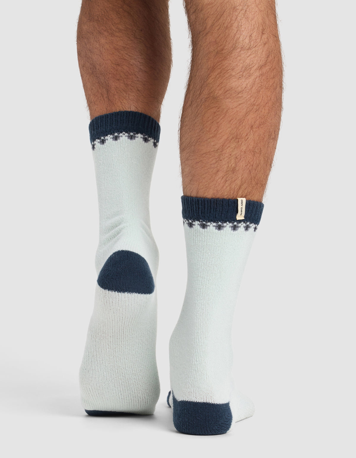 A person stands on tiptoe, viewed from behind, wearing Cozy Earth’s The Mistletoe Plush Lounge Sock—light-colored with dark blue heels, toes, cuffs, and a dotted pattern near the top.