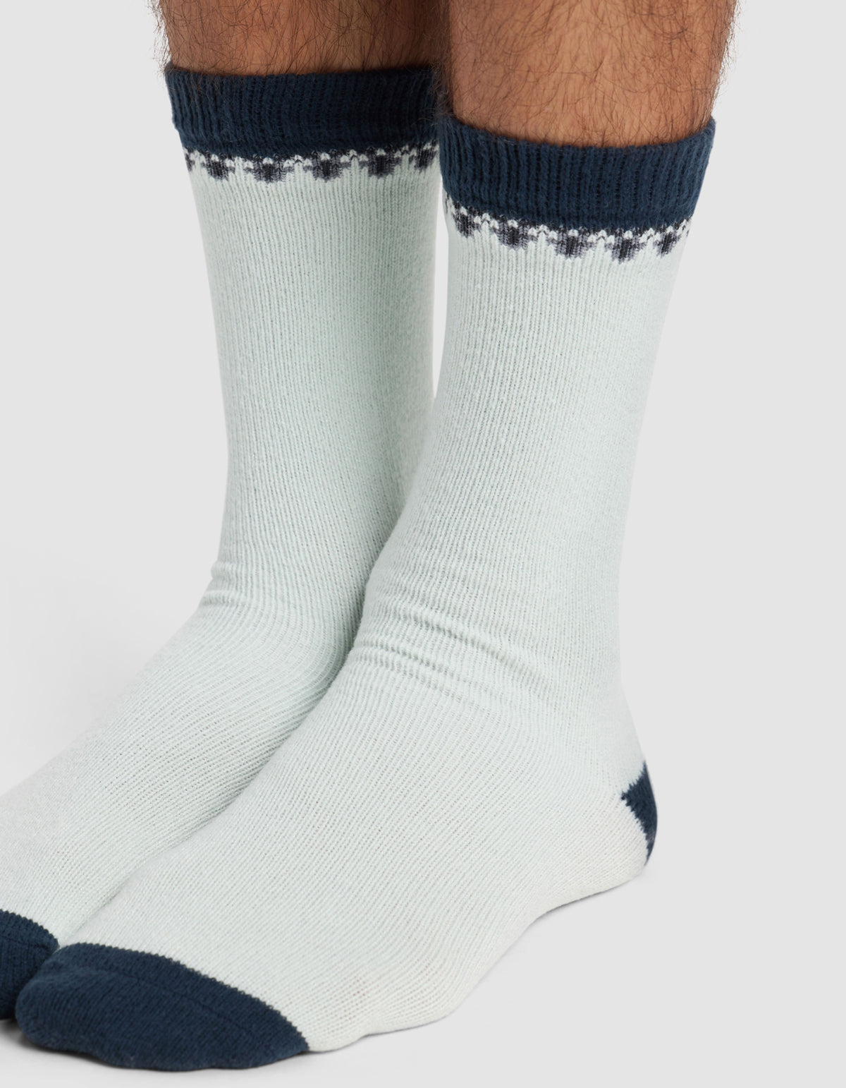 A pair of feet wear The Mistletoe Plush Lounge Sock by Cozy Earth, featuring light fabric with dark blue toes, heels, and cuffs. The cuffs display a decorative dark blue pattern. The background is plain and light-colored.