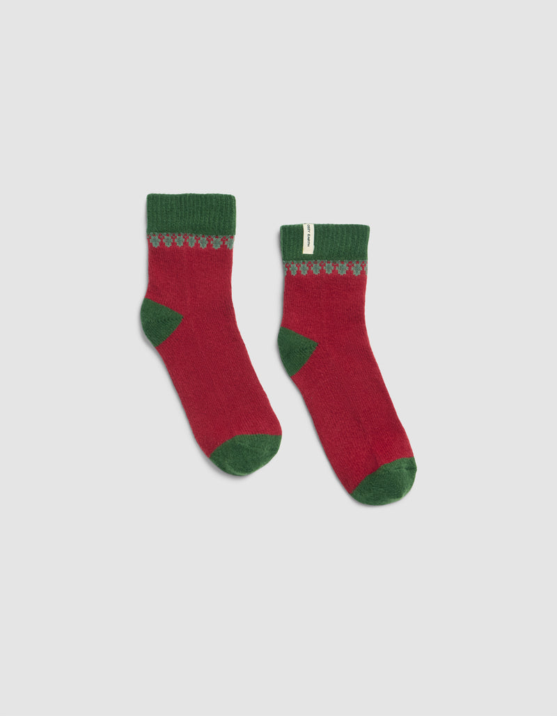 The Mistletoe Plush Modern Crew Sock by Cozy Earth features red fabric with green cuffs, heels, and toes, plus a decorative green and gray pattern on top. They are displayed flat against a plain white background.