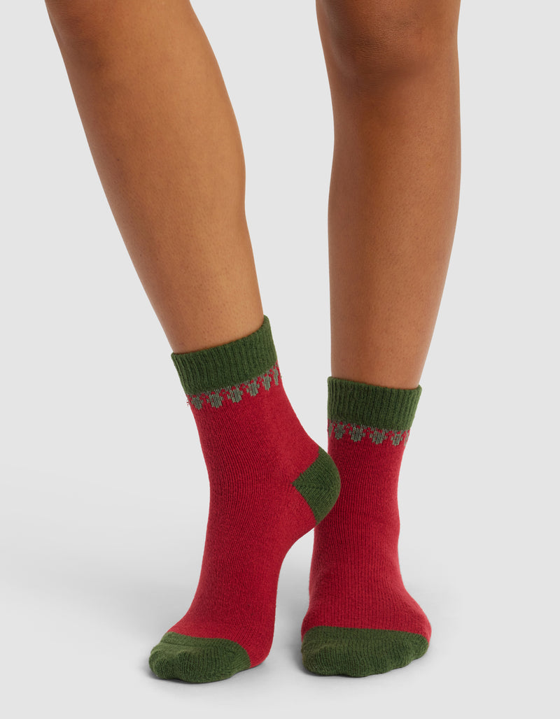 A person wearing The Mistletoe Plush Modern Crew Sock by Cozy Earth, featuring red with green heels, toes, and cuffs accented by a green and gray decorative pattern. Background is plain white.