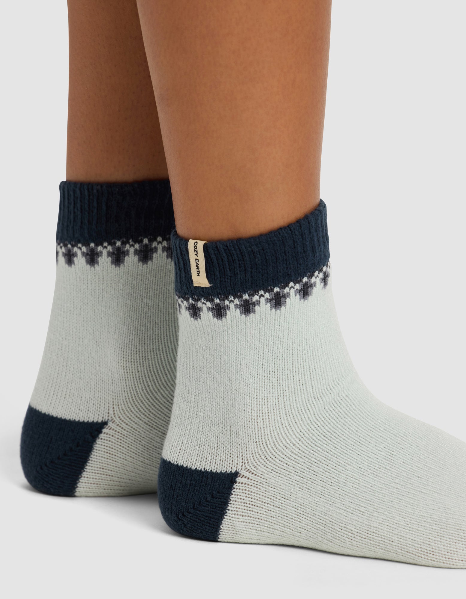 Close-up of two feet wearing Cozy Earth’s The Mistletoe Plush Modern Crew Socks—thick, light-colored with dark heels, toes, and cuffs, plus a pattern near the top and a small beige tag on one cuff.|Color:Mist