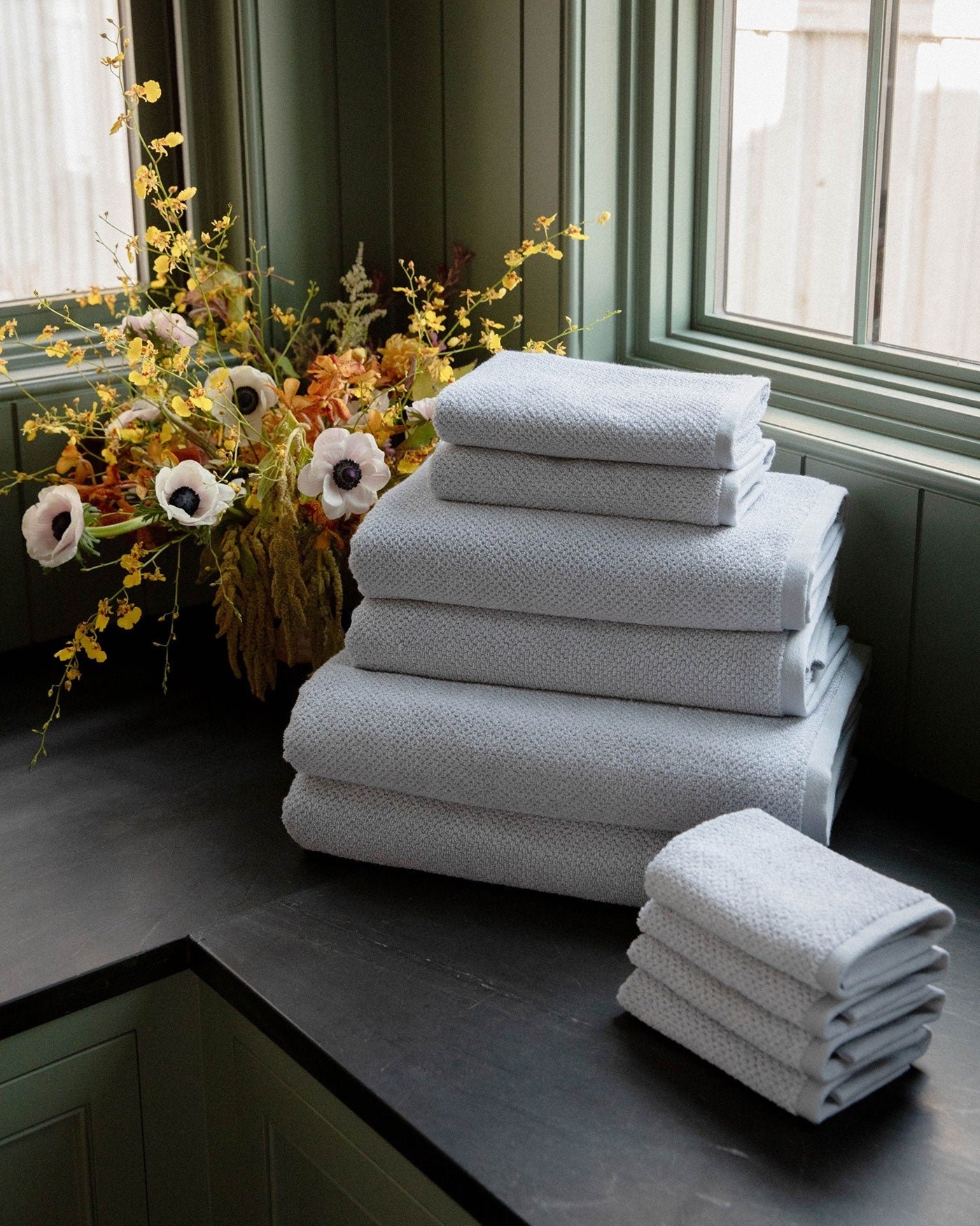 The Complete Nantucket Bath Bundle by Cozy Earth is neatly stacked on a dark countertop near a window, with a vase of yellow and white flowers in the background.|Color:Heathered Harbor Mist
