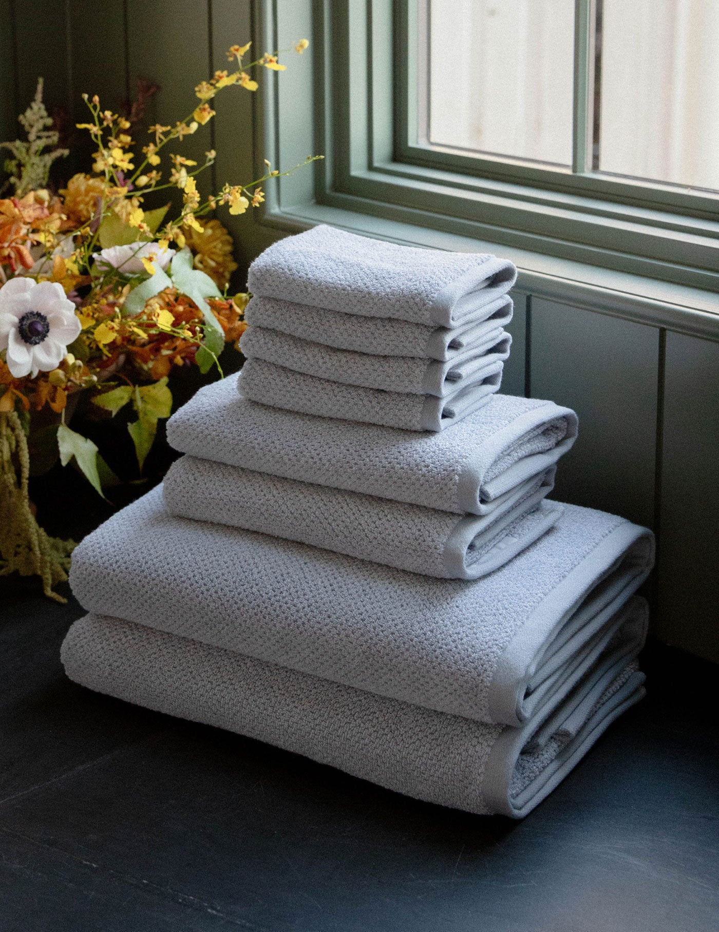 The Cozy Earth Nantucket Bath Towel Set, in varying sizes, is neatly stacked on a dark surface near a window, with yellow and white flowers in the background. 