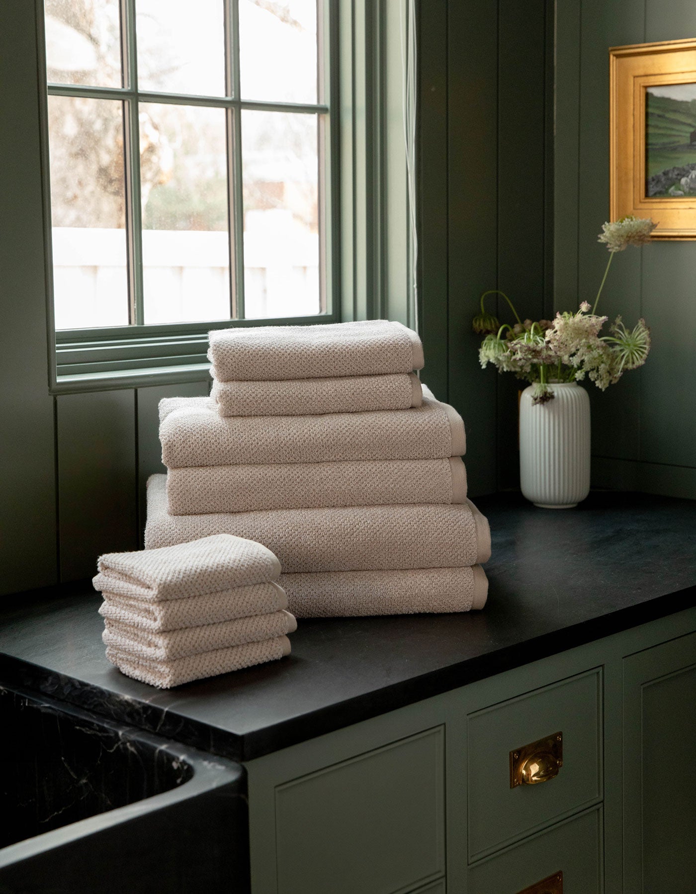 The Complete Nantucket Bath Bundle by Cozy Earth, in beige, is neatly stacked on a black countertop near a window with a vase of flowers and pale green cabinetry in the background.|Color:Heathered Sand