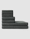 The Nantucket Bath Towel Set by Cozy Earth features dark gray towels in various sizes, neatly stacked in a step-like pattern against a plain light gray background.|Color:Graphite
