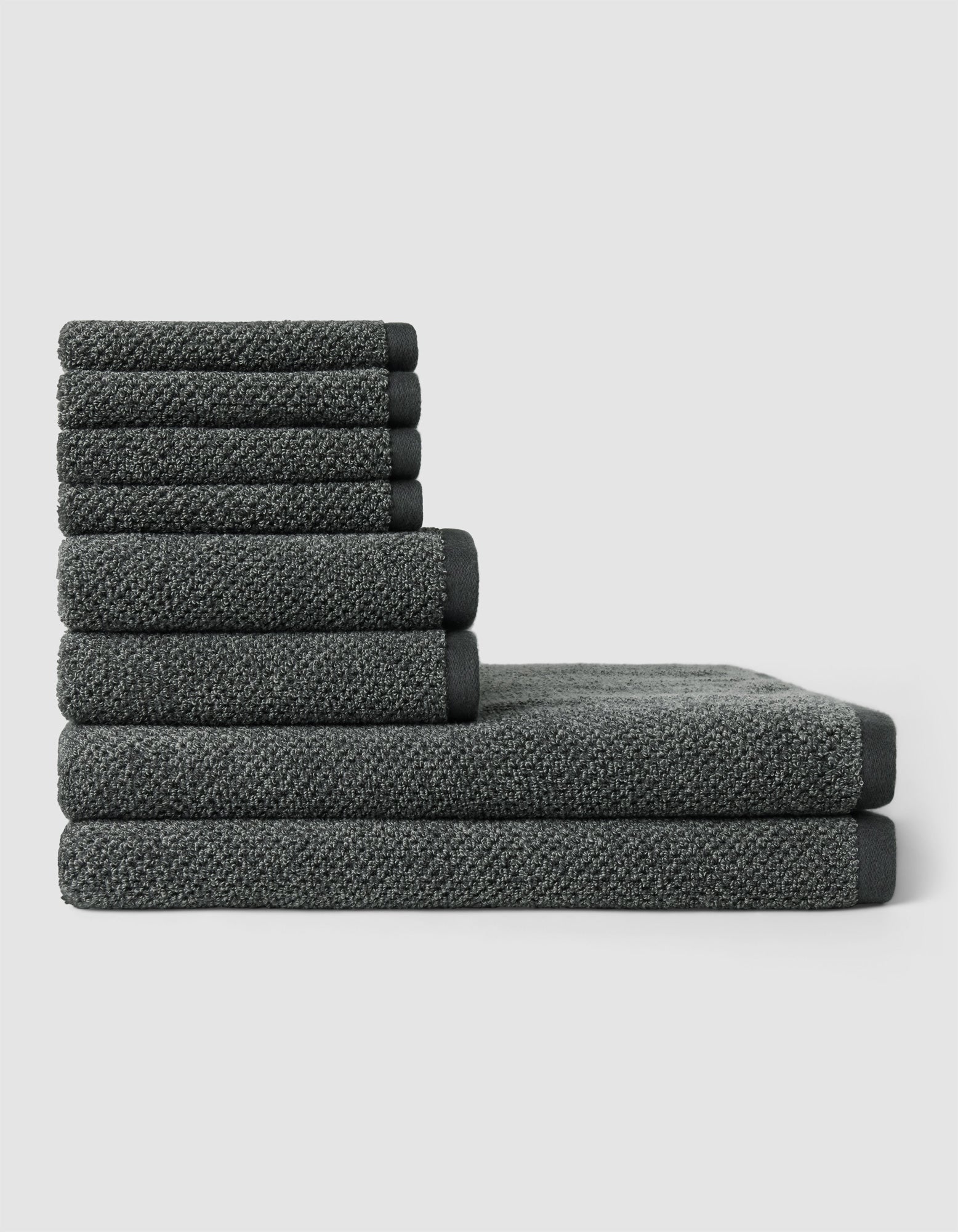 The Nantucket Bath Towel Set by Cozy Earth features dark gray towels in various sizes, neatly stacked in a step-like pattern against a plain light gray background.