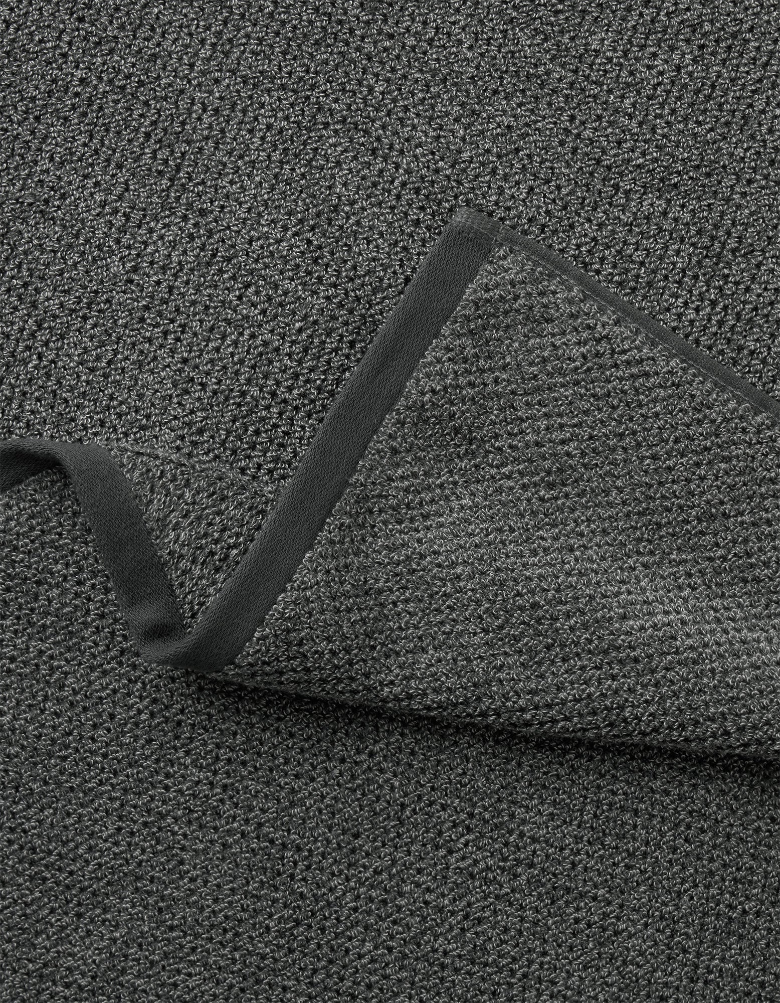 A close-up of Cozy Earth's Nantucket Bath Towel Set shows its textured dark gray fabric with a folded corner, revealing the same woven material on both sides and a smooth, dark border along the edge.|Color:Graphite