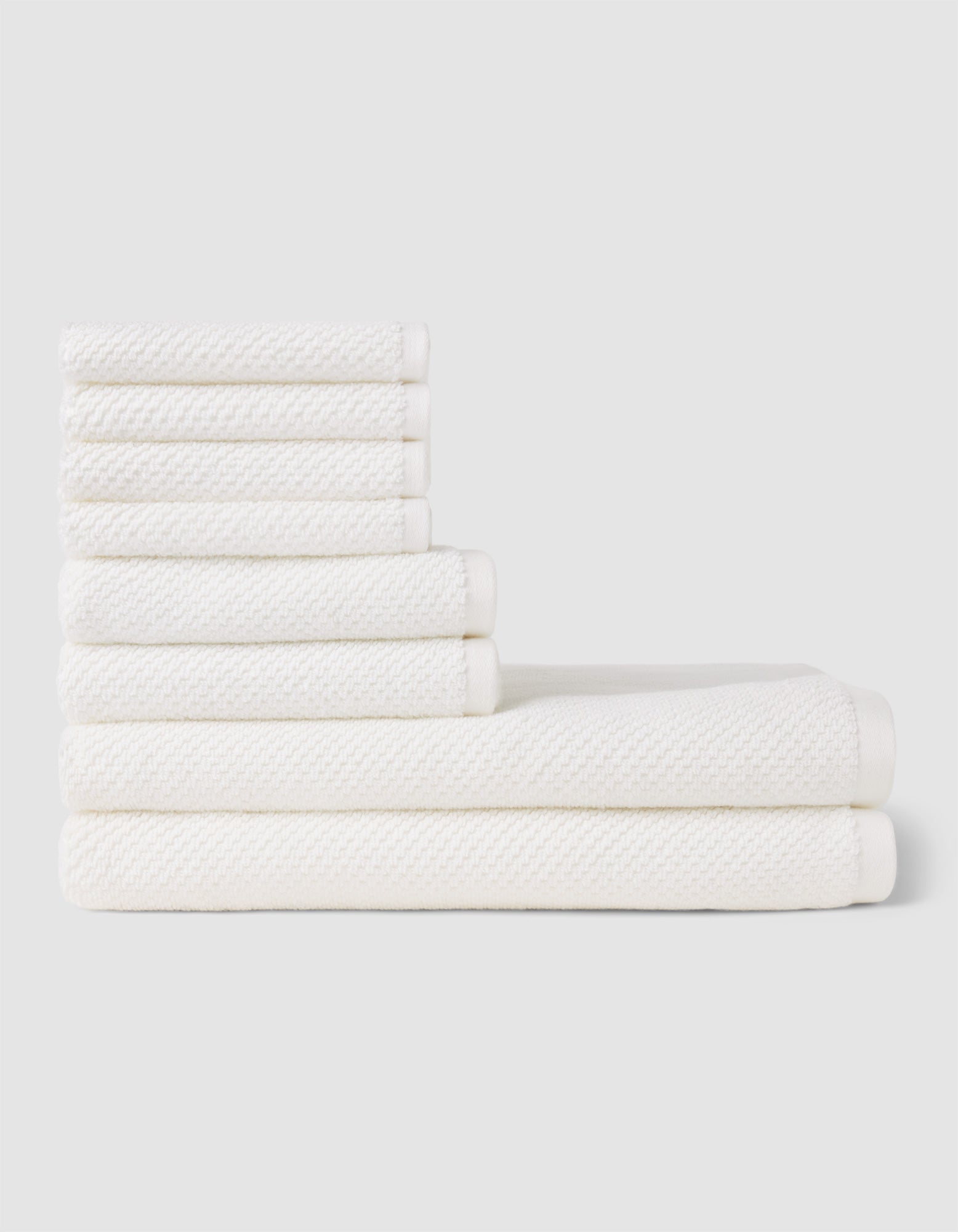The Cozy Earth Nantucket Bath Towel Set features seven white textured towels—three large and four small—neatly stacked in two rows on a light gray background, showcasing their soft, plush feel.