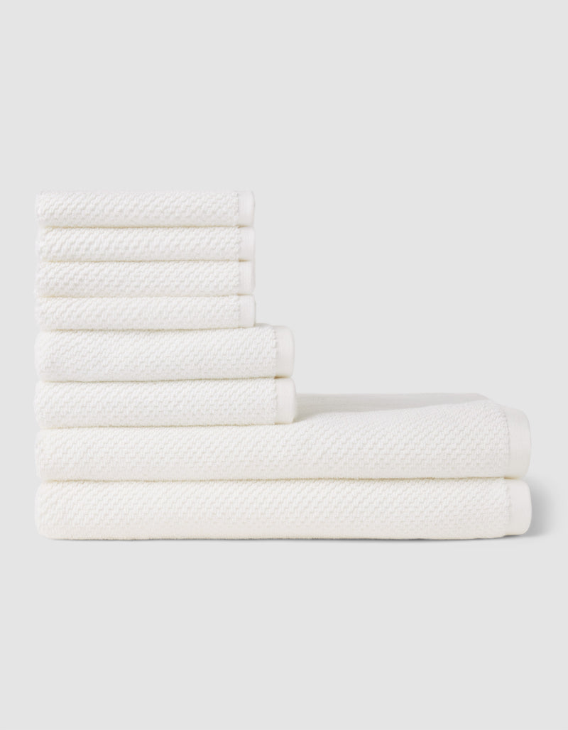 The Cozy Earth Nantucket Bath Towel Set features seven white textured towels—three large and four small—neatly stacked in two rows on a light gray background, showcasing their soft, plush feel.