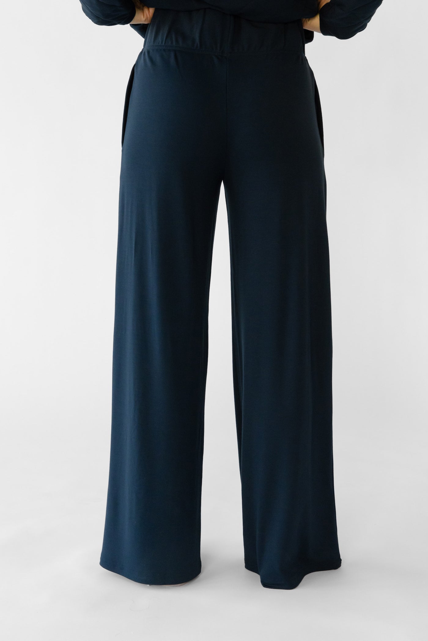 A person stands with hands on hips, wearing Cozy Earth's Women's Ultra-Soft Bamboo Wide Leg Pull On Pant & Hoodie Set in dark blue against a plain light gray background. The pants have a wide-leg cut and appear very comfortable. 