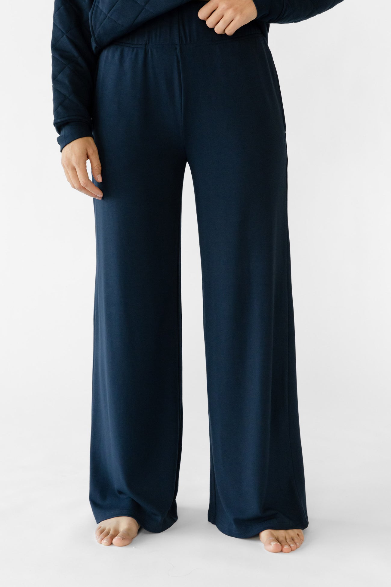 A person wears Cozy Earth's Women's Ultra-Soft Bamboo Wide Leg Pull-On Pant in dark blue and a matching long-sleeved top, standing barefoot on a plain white background with hands resting casually by their sides. |Color:Navy