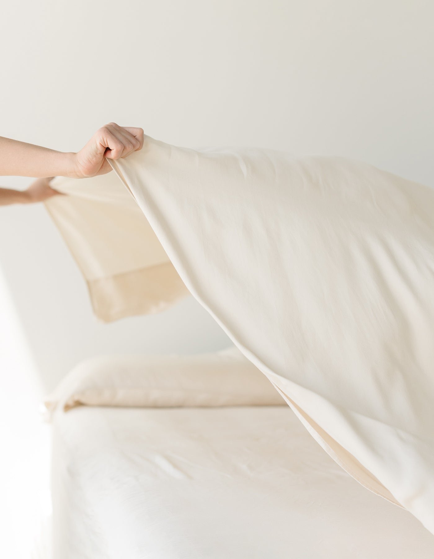 A person is making a bed with Cozy Earth's Bamboo Sheet Set in beige. The room is bright and minimalist, with soft natural light illuminating the bed and a partially visible pillow in the background. 