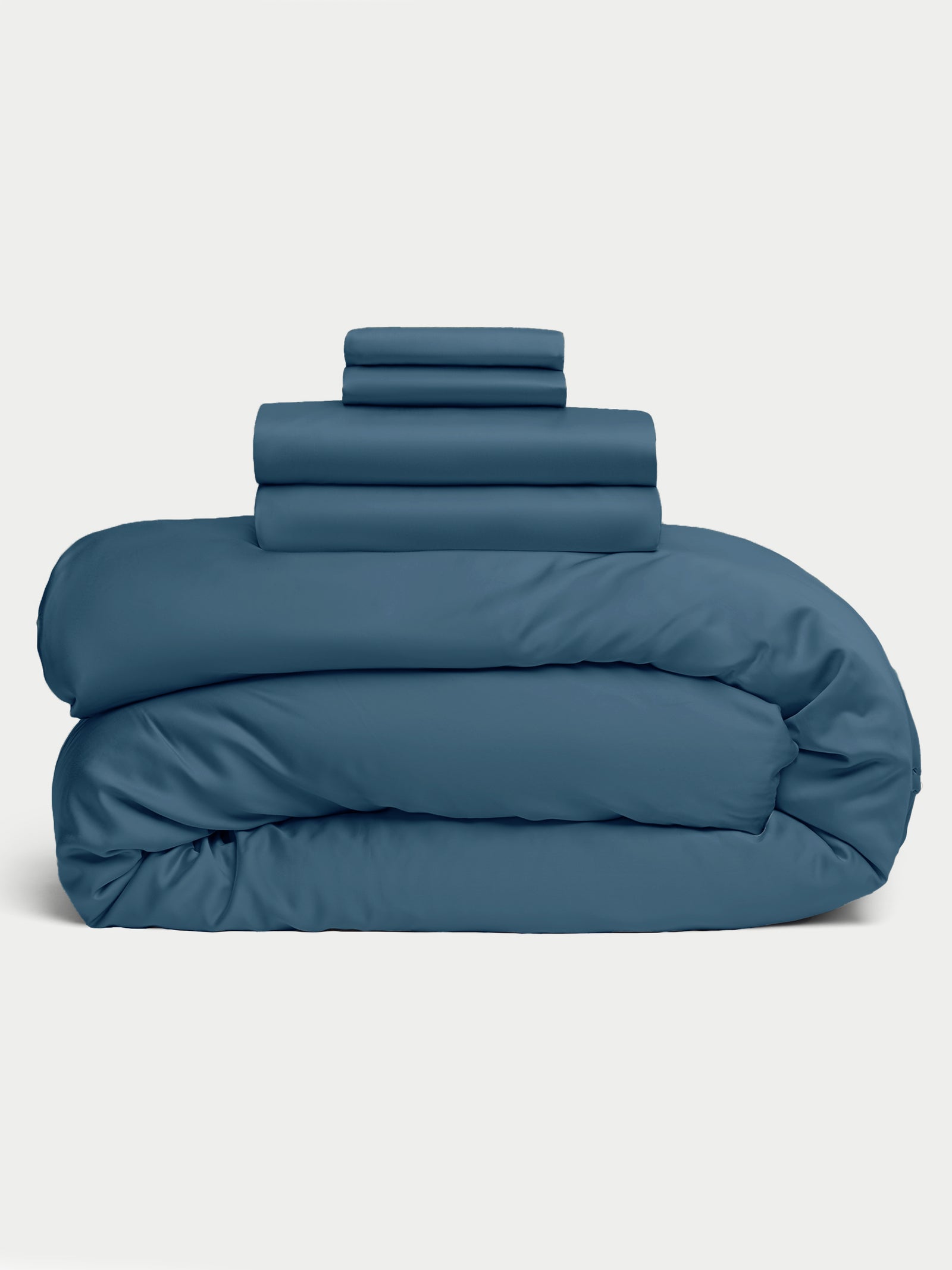 The Bamboo Bedding Deluxe Bundle by Cozy Earth is neatly stacked, featuring two folded pillowcases, a fitted sheet, and a flat sheet in dark teal on a fluffy comforter against a pale background. 