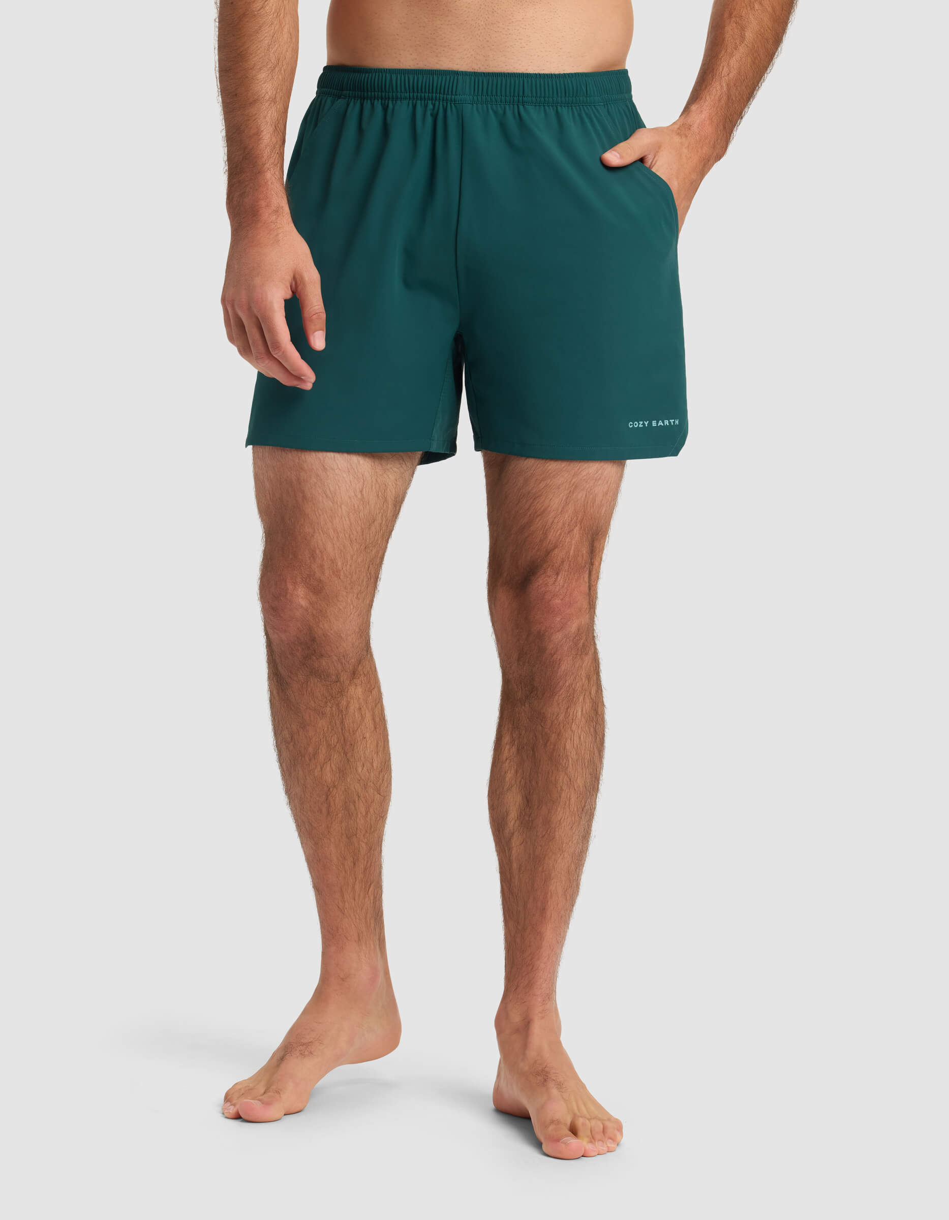 A barefoot man stands against a plain background, wearing Cozy Earth Men's Day & Night Short 5" in teal with an elastic waistband and one hand in his pocket.