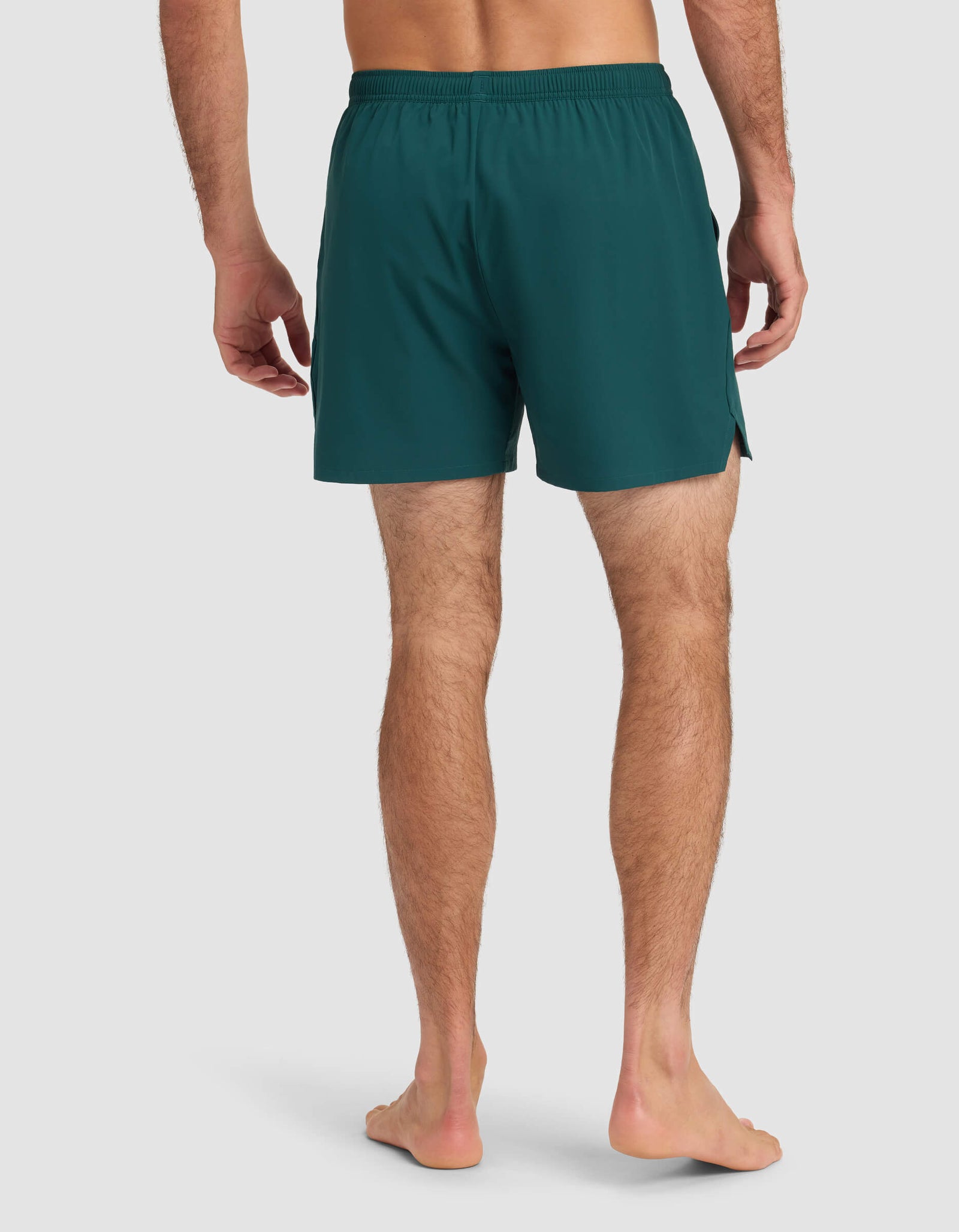 A barefoot person stands on a light background, shown from behind, wearing Cozy Earth Men's Day & Night Short 5" in dark teal with an elastic waistband and side slits.