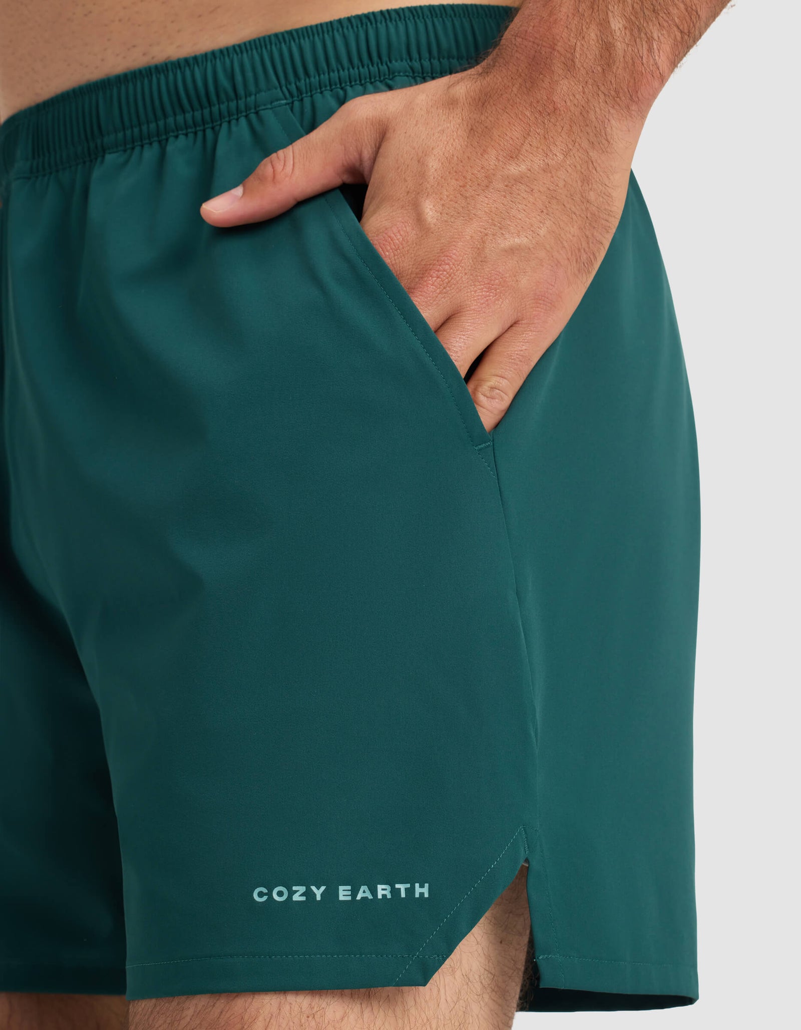 Close-up of a person wearing Cozy Earth's Men's Day & Night Short 5" in teal, with their right hand in the pocket. "COZY EARTH" is printed near the hem against a plain light gray background.