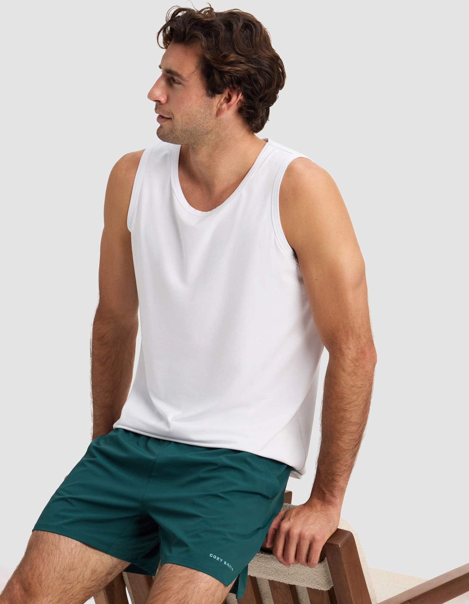 A man wearing a white sleeveless tank top and Cozy Earth Men's Day & Night Short 5" in dark green sits on a wooden bench, looking to his left against a plain light background.