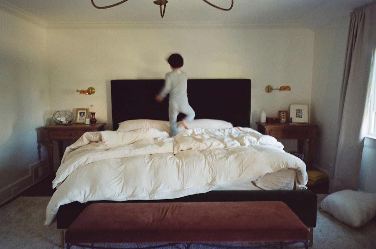 Person standing on a bed in a bedroom with a chandelier and side tables.