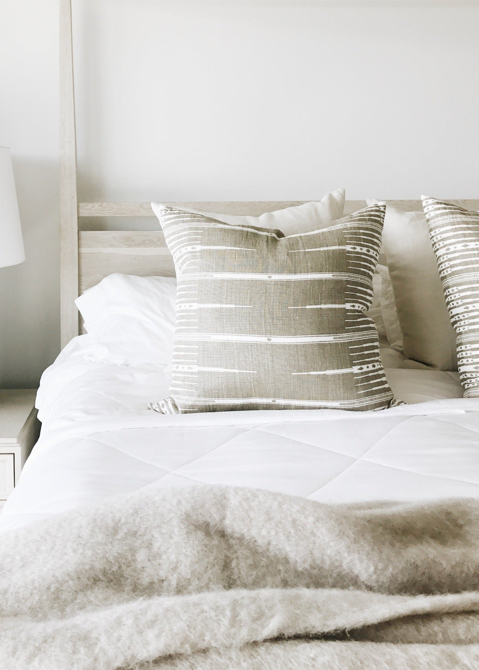 A neatly made bed features the Cozy Earth Bamboo Viscose Comforter in white and beige, topped with two patterned throw pillows and a soft blanket. A light nightstand holds a white lamp with dark beads, dish, candle, and decor item.