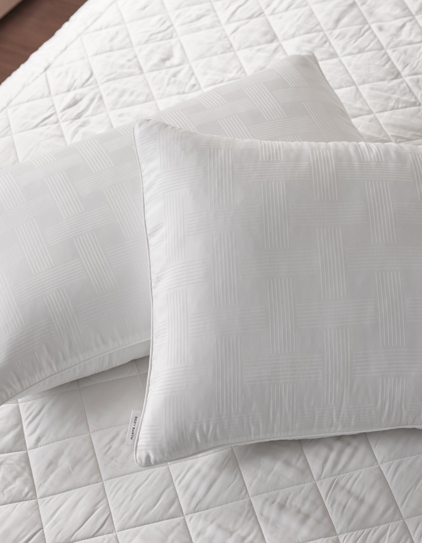 Two Cozy Earth Pillow Protectors in white, featuring a subtle geometric pattern, rest on a matching quilted bedspread. The bedding looks clean, crisp, and neatly arranged.