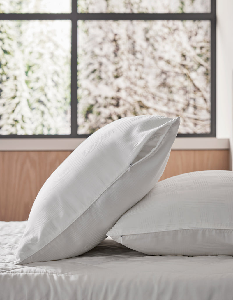 Two Cozy Earth Pillow Protectors in white are stacked on a neatly made bed with a wooden headboard. A snowy tree view from the window behind creates a bright, peaceful atmosphere.