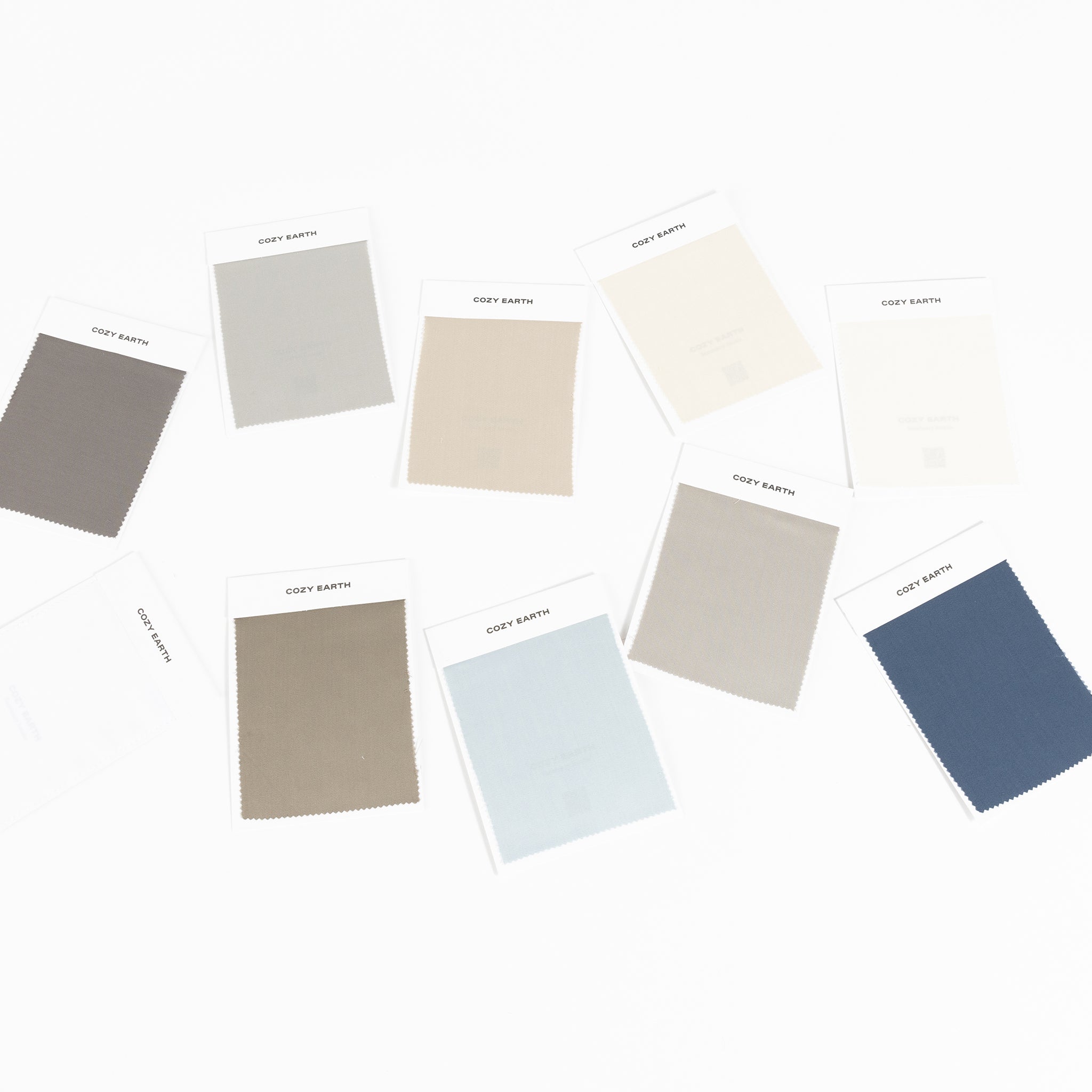 A selection of Bamboo Sheet Swatches from Cozy Earth arranged on a plain surface, showcasing a variety of shades in grey, beige, blue, and green. Each swatch is affixed to a labeled card.