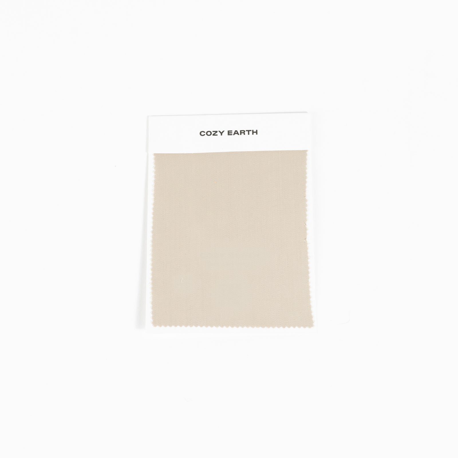 A square fabric sample from Cozy Earth, identified as the Bamboo Sheet Swatch. The swatch is a simple beige color with a zigzag edge design and is attached to a white card. 