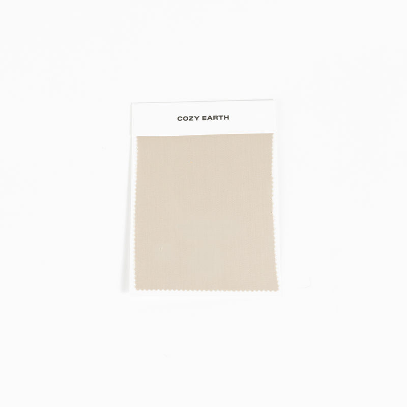 A square fabric sample from Cozy Earth, identified as the Bamboo Sheet Swatch. The swatch is a simple beige color with a zigzag edge design and is attached to a white card.