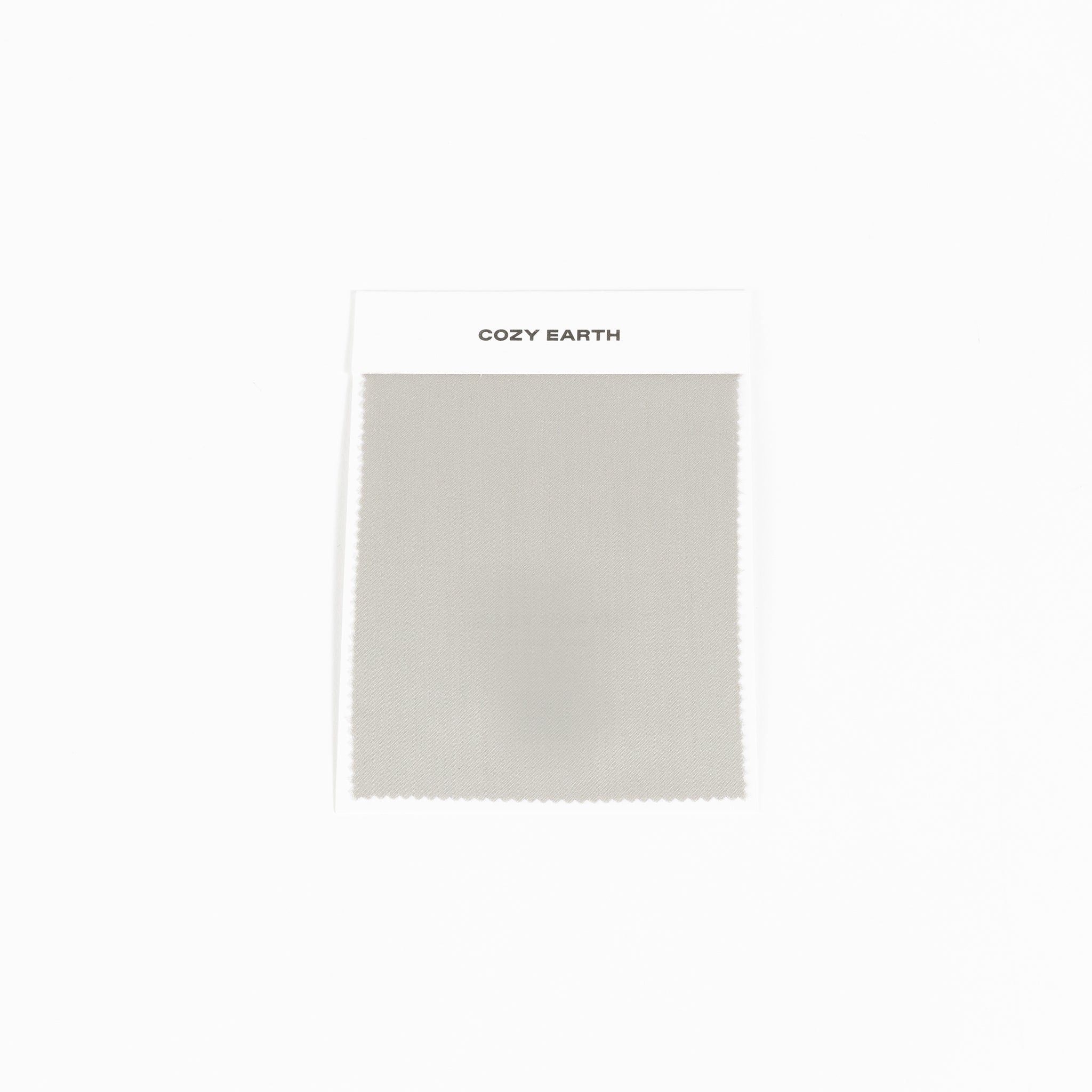 A "Bamboo Sheet Swatch" from Cozy Earth, featuring a light gray fabric with a smooth texture and a zigzag edge, displayed against a white background. |Color:Light Grey