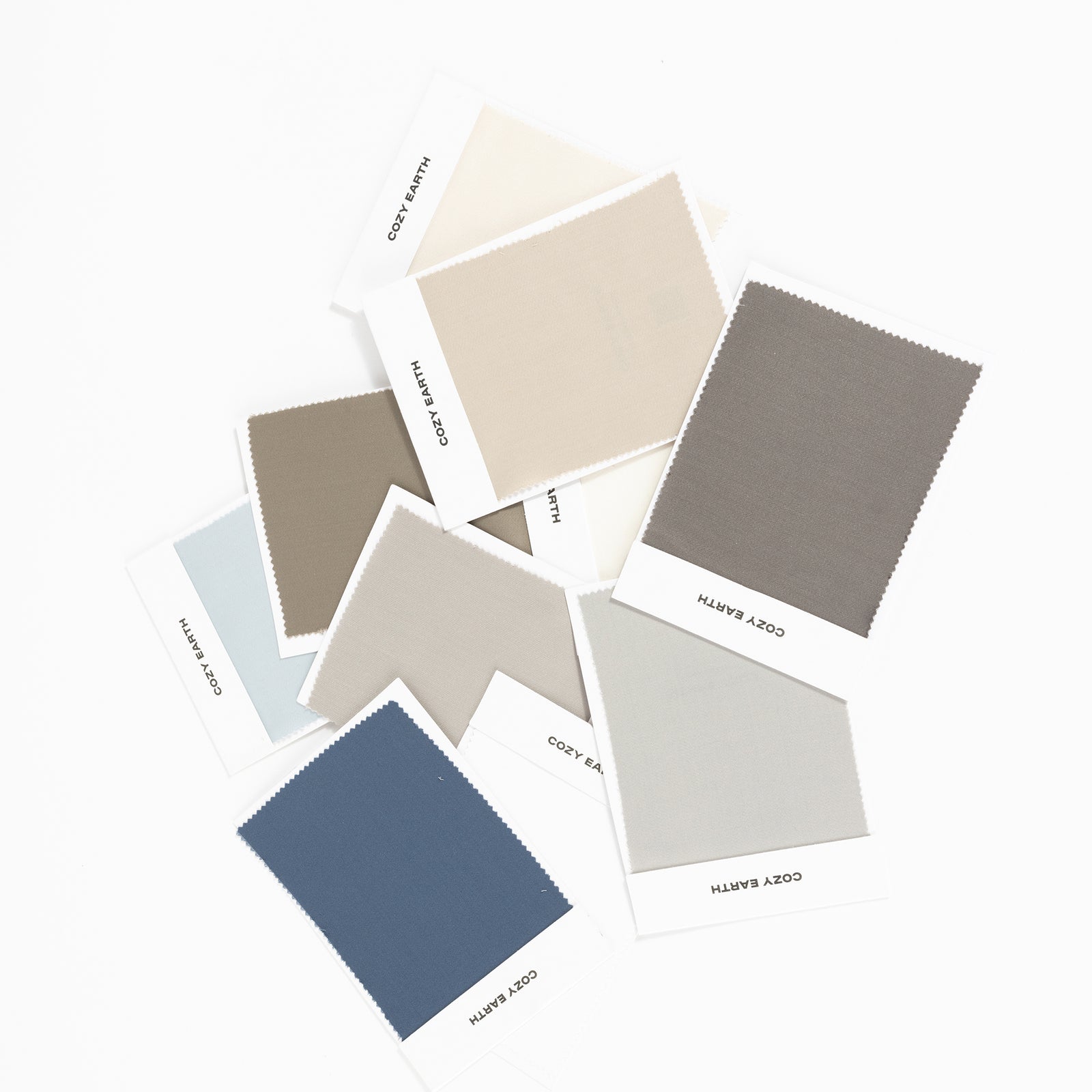 A variety of "Cozy Earth" Bamboo Sheet Swatches are laid out on a white surface, showcasing an array of colors including driftwood, oat, walnut, charcoal, light grey, dove grey, pacific blue, shore, creme, and white