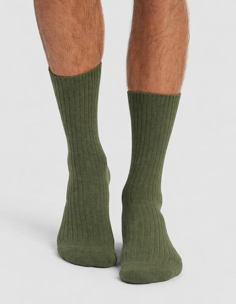 A person wears Cozy Earth's Men's Plush Lounge Calf Sock in ribbed olive green, standing on a light gray surface with the socks shown from back and side angles.