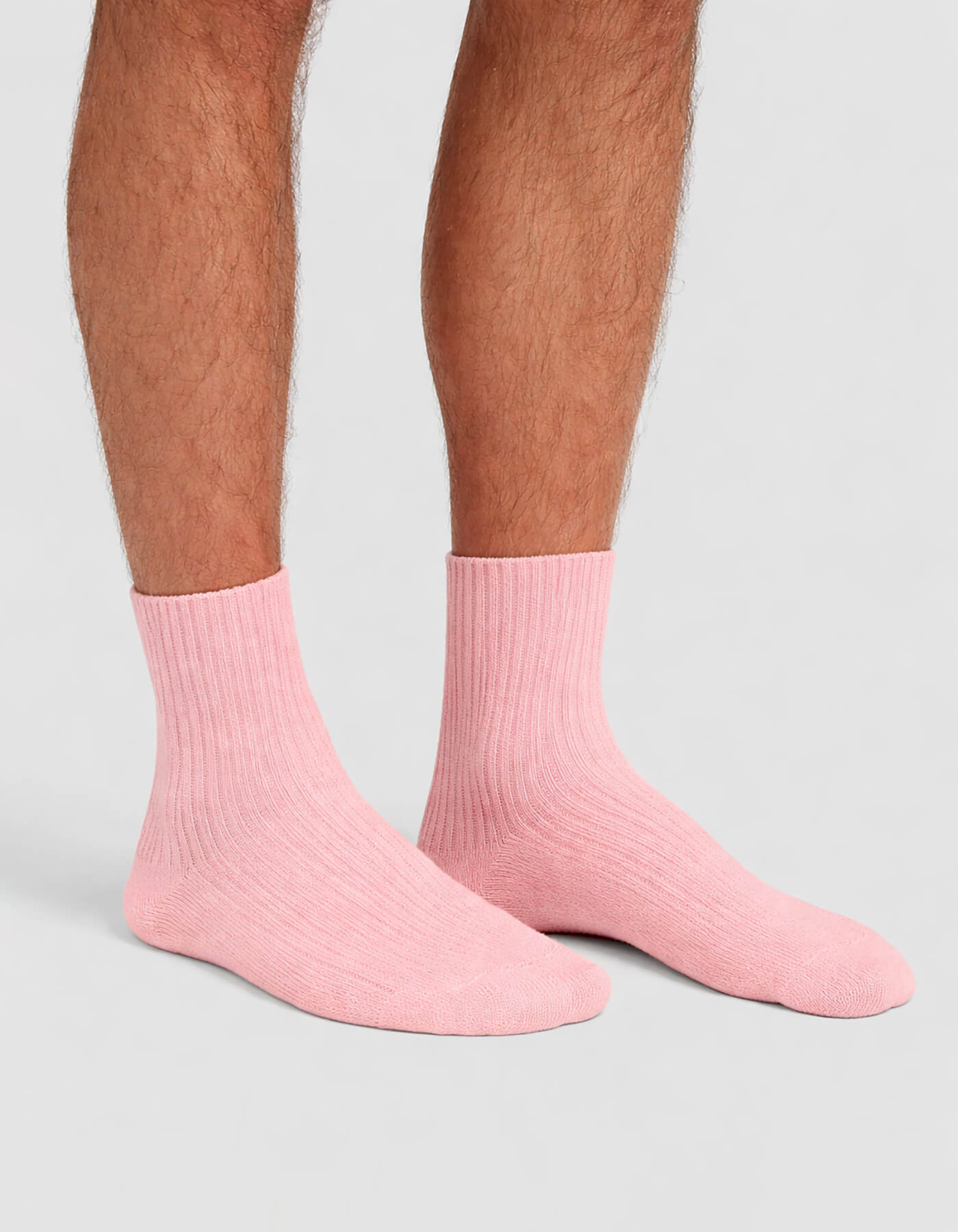 A person wears Cozy Earth Men's Plush Lounge Crew Socks in light pink, standing on a light gray surface. Only the lower legs and feet are visible.