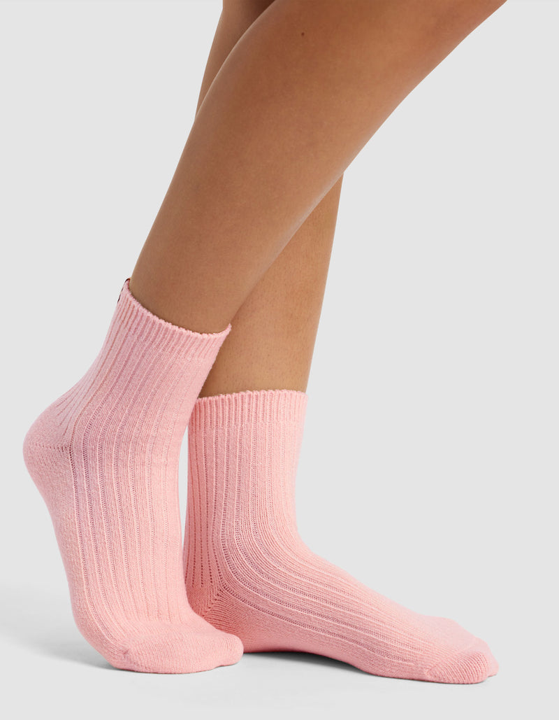 A pair of feet wears The Plush Modern Crew Sock by Cozy Earth in light pink, featuring a ribbed, cozy texture against a plain light gray background.
