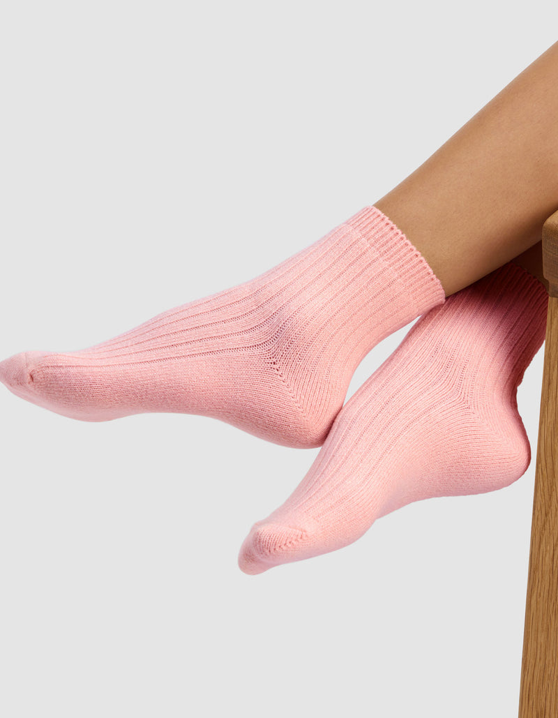 A person wearing The Plush Modern Crew Sock by Cozy Earth in light pink rests their feet on a wooden surface against a plain light gray background.
