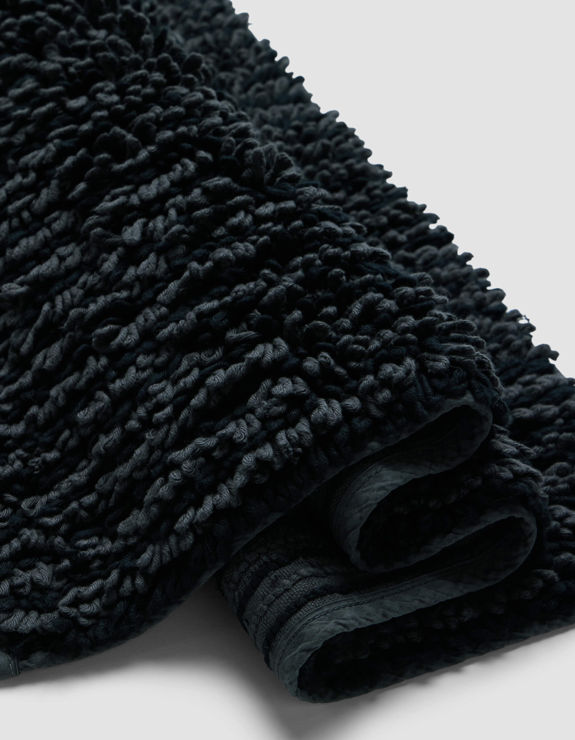 A close-up of the Plush Loop Bath Mat by Cozy Earth shows its dark, textured fabric with a thick, looped pile and soft, plush folds against a plain light background.|Color:Graphite