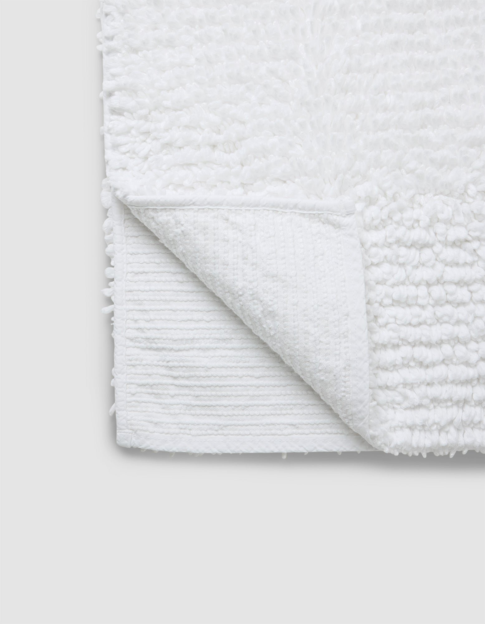 A close-up of the Cozy Earth Plush Loop Bath Mat in white, featuring a soft, textured surface with one corner flipped to show its flat underside. The mat is set against a plain light gray background.