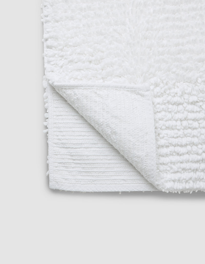A close-up of the Cozy Earth Plush Loop Bath Mat in white, featuring a soft, textured surface with one corner flipped to show its flat underside. The mat is set against a plain light gray background.