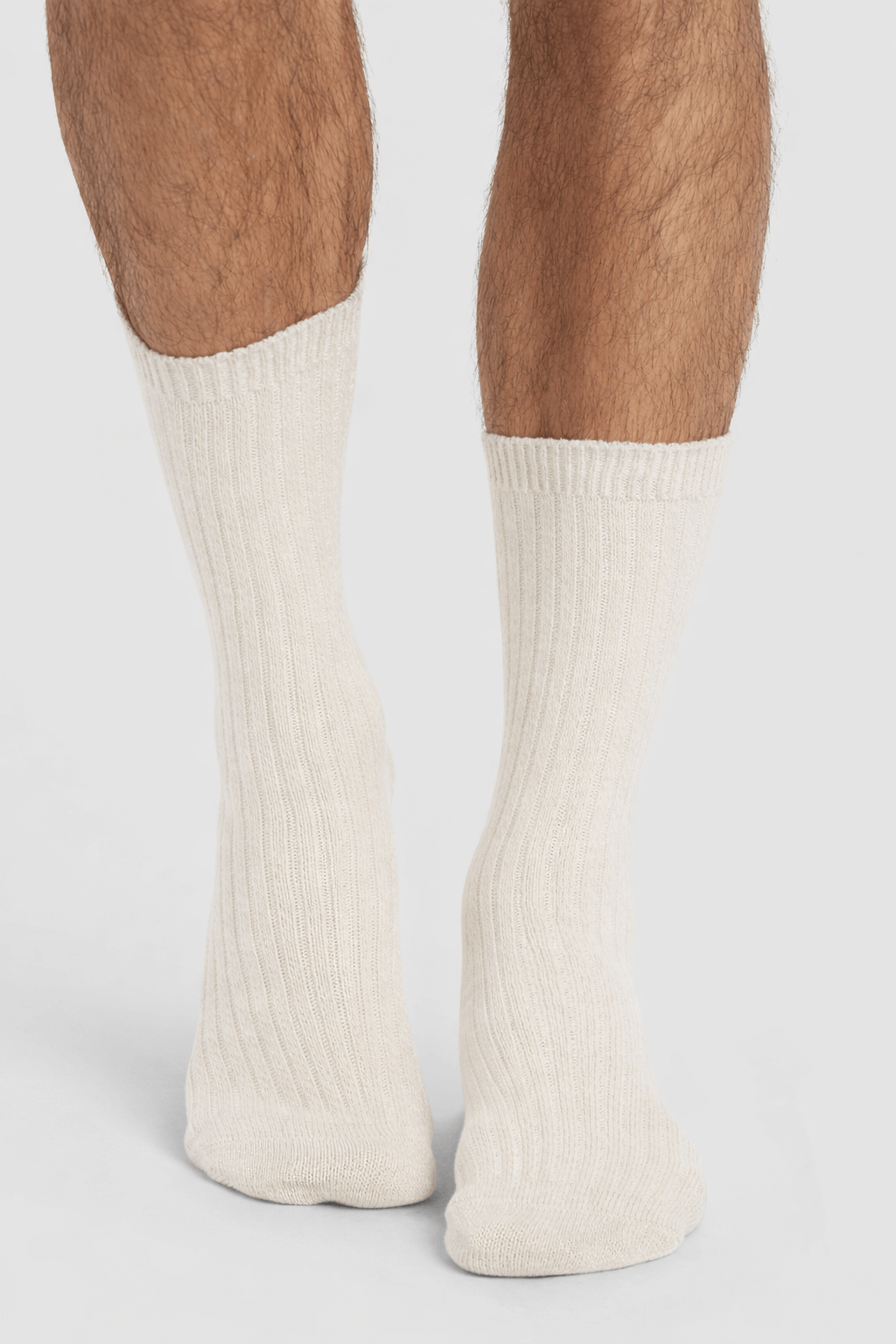 A close-up of a person's lower legs wearing Cozy Earth's Men's Plush Lounge Quarter Sock 3-Pack in cream, ribbed design, standing on a white surface.