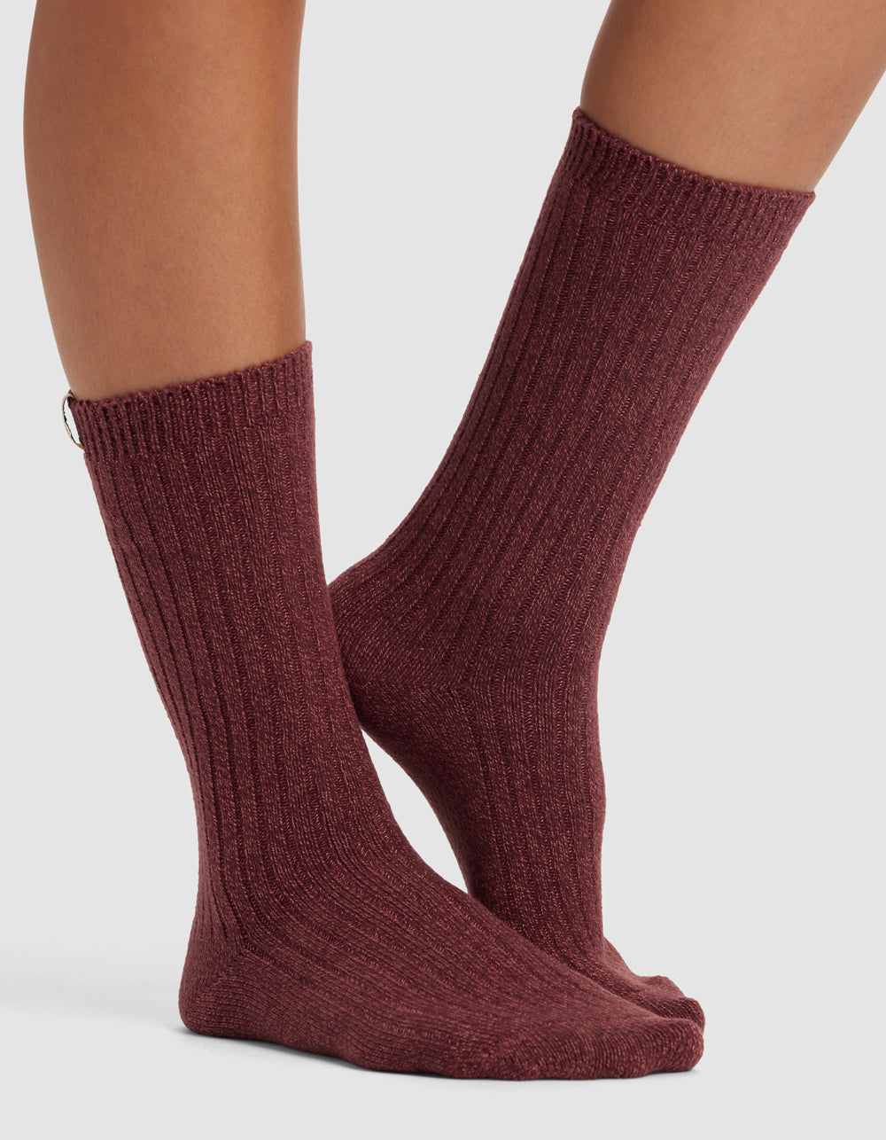 A pair of bare legs wears The Plush Lounge Calf Sock from Cozy Earth in ribbed, dark mauve, mid-calf length, featuring a snug fit and subtle texture, standing on a light surface.