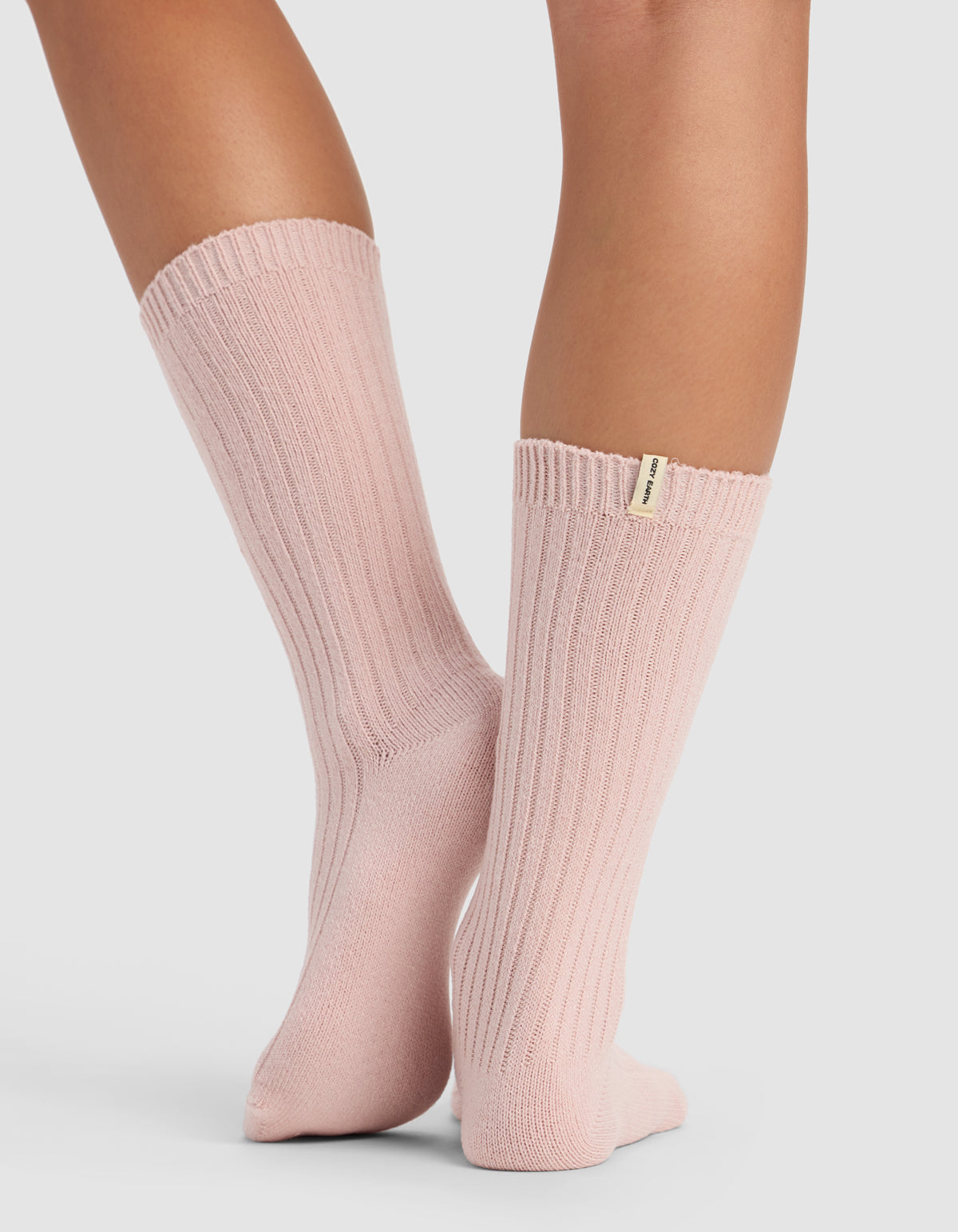 A person models Cozy Earth’s The Plush Lounge Calf Sock 3-Pack in light pink ribbed style, standing on a light surface with ankles crossed. A small fabric tag is visible near the top of one sock.