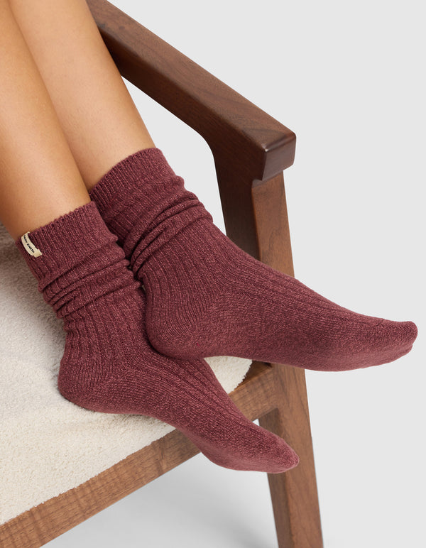 A person sits on a cushioned wooden chair with crossed legs, wearing Cozy Earth’s Plush Lounge Socks in maroon from the 3-Pack. The textured socks are slightly slouched. The background is plain and light-colored.|Color:Burgundy/Rouge/Dusty Rose