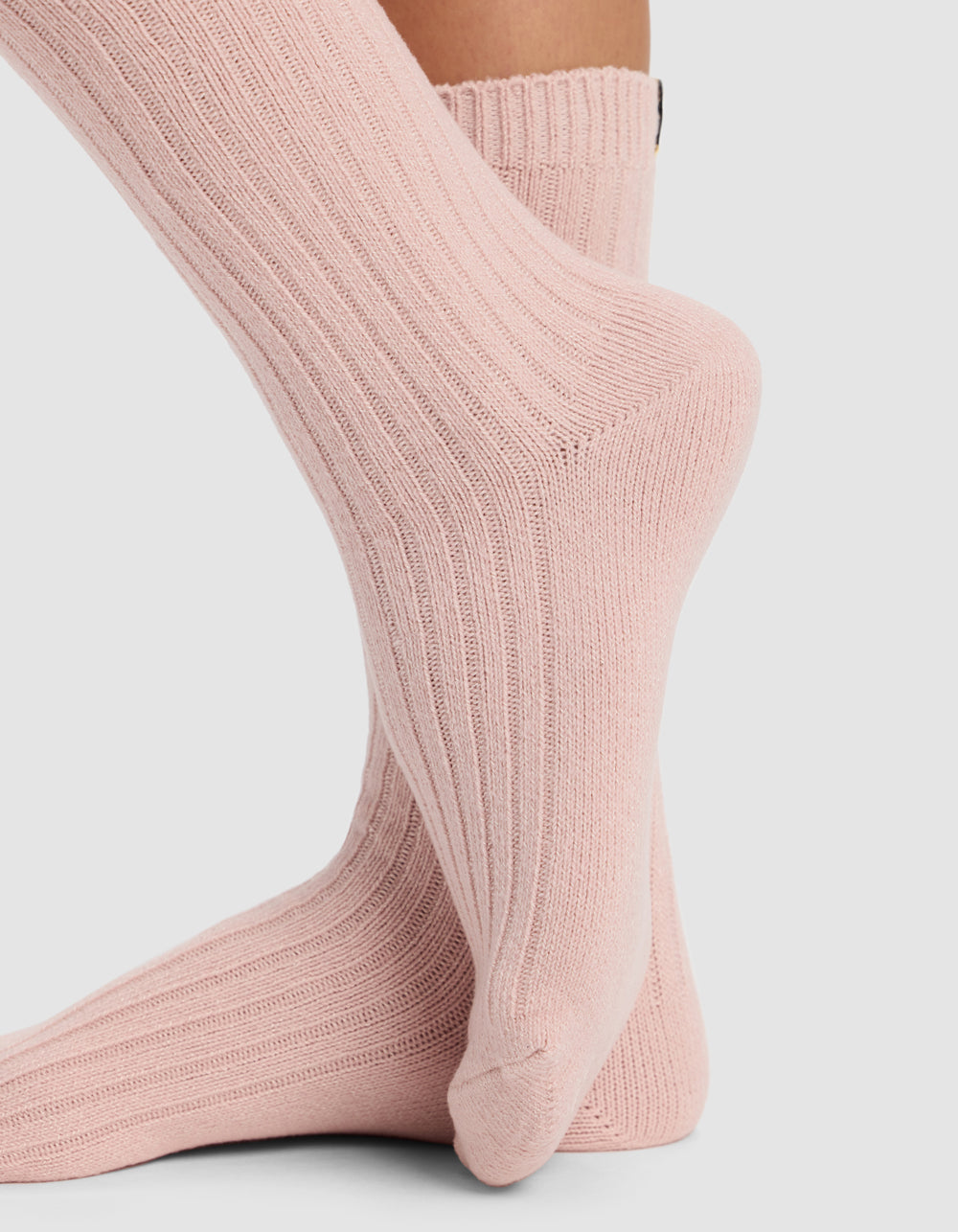 Close-up of two feet wearing light pink Cozy Earth The Plush Lounge Calf Sock 3-Pack socks. One foot is flat on a plain light gray background, while the other is elevated, resting on the toes.
