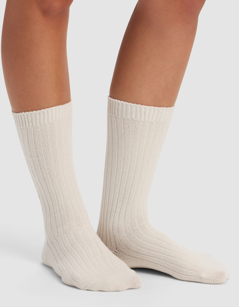 Someone wearing Cozy Earth's The Plush Lounge Calf Sock 3-Pack in a thick, cream-colored ribbed style that reaches mid-calf, standing on a white surface with only their lower legs and feet visible.