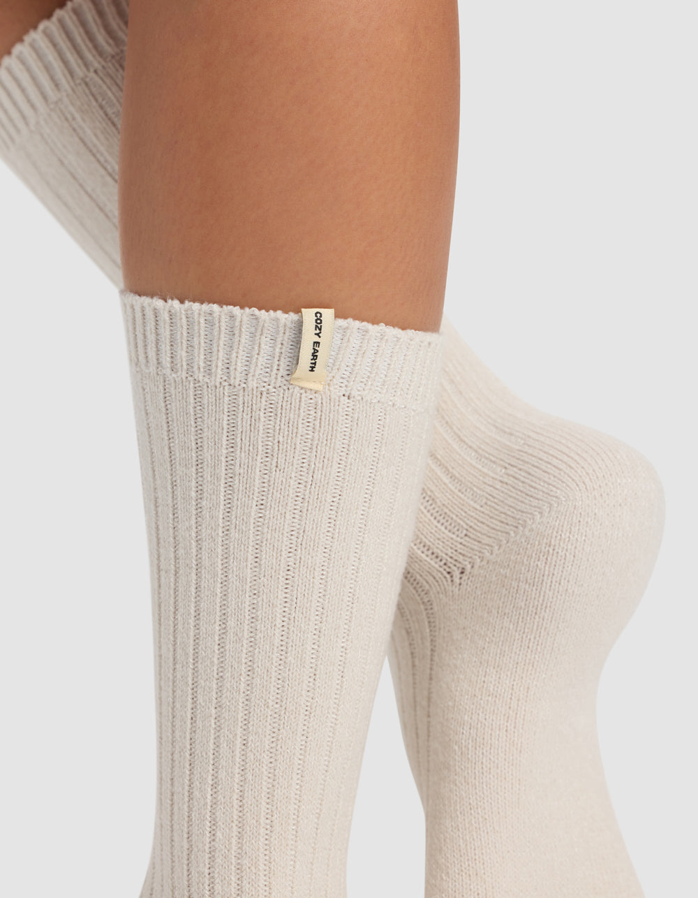 Close-up of crossed legs wearing thick, ribbed, cream-colored socks from the Plush Lounge Calf Sock 3-Pack by Cozy Earth. A small cream tag with black “COZY EARTH” text is on the ribbed cuff. Background is plain white.