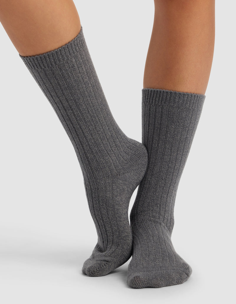 Someone wearing Cozy Earth’s Plush Lounge Calf Sock 3-Pack in dark gray stands on a white surface with one foot crossed over the other.